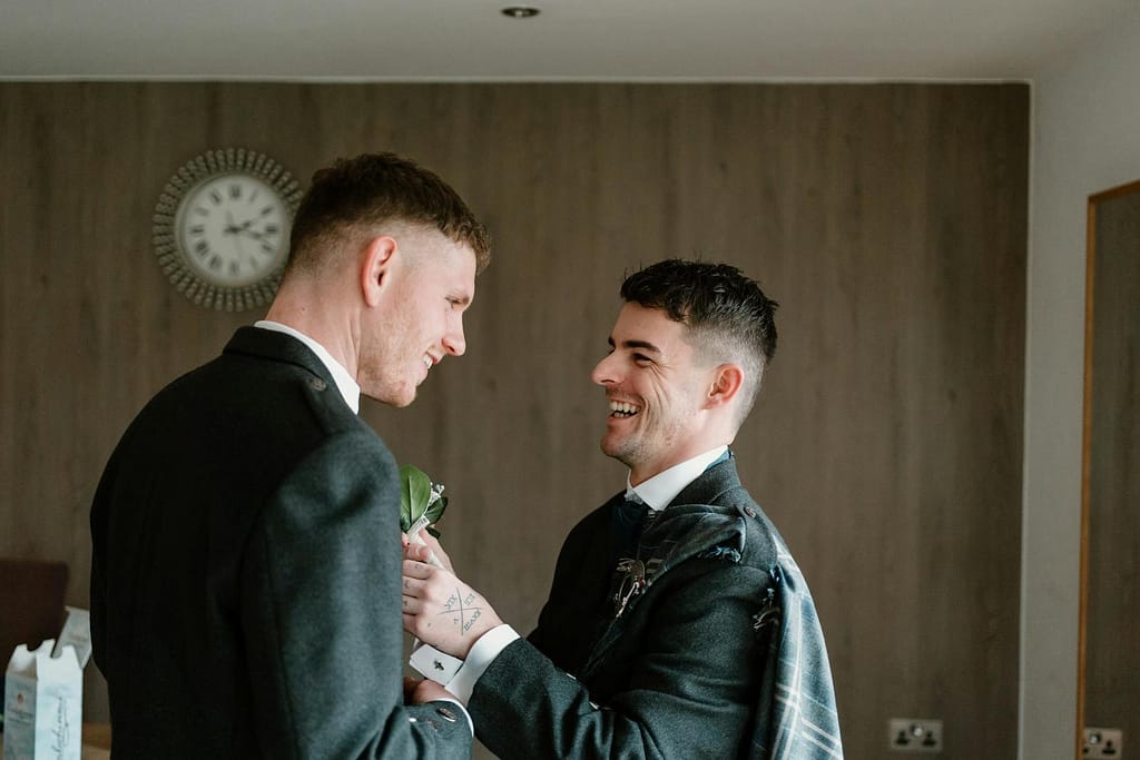 Two men in suits, joyfully preparing for a wedding indoors, expressing happiness and camaraderie.