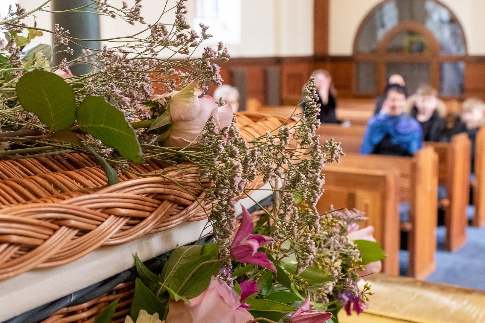 Willow coffin decorated with flowers and mourners seated in wooden pews