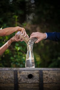 Unrecognizable groom and bride pouring sand from jars into glass vase while performing wedding ritual on street during holiday celebration on blurred background