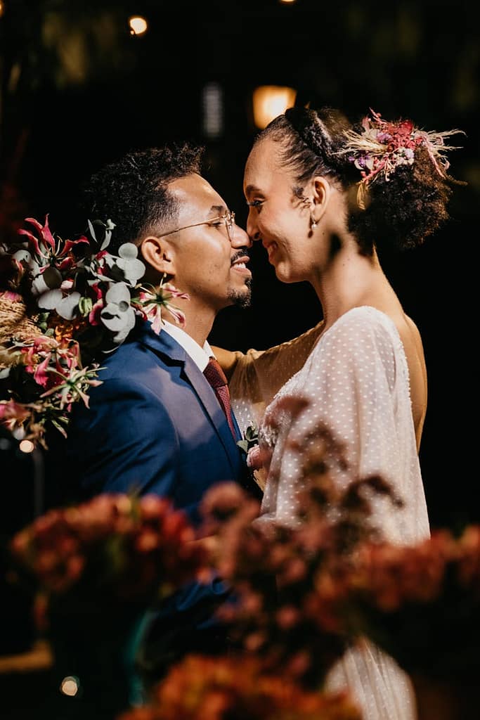 Side view of young smiling bride embracing content black groom in suit while looking at each other near flower decor