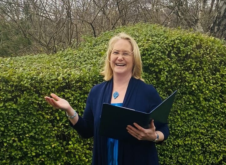 Fleur laughing with one hand holding a folder and the other outstretched in a garden ceremony with a hedge behind her