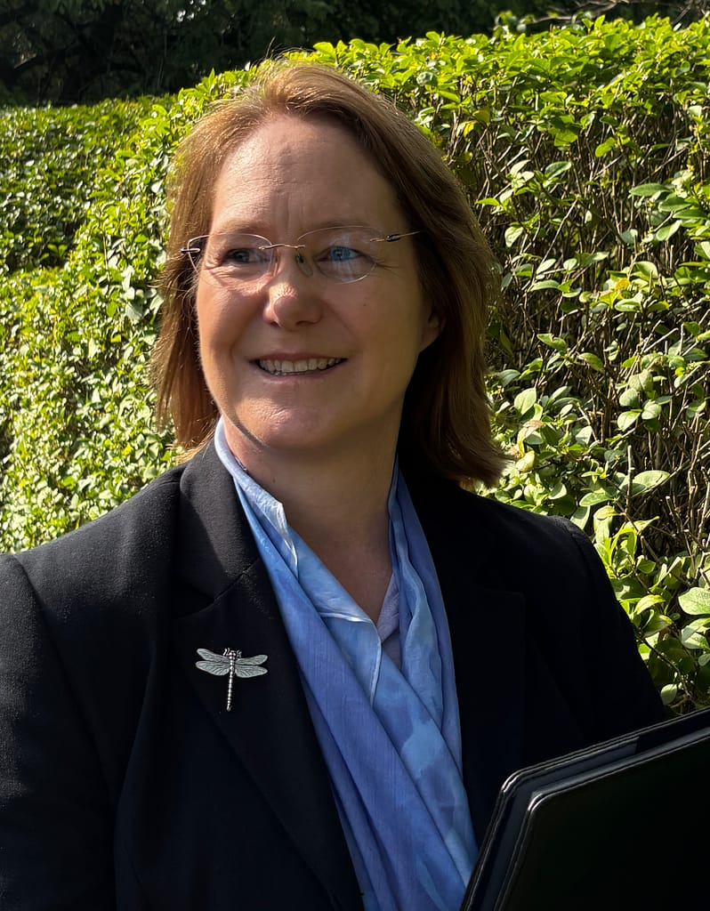 Fleur smiling and holding a folder, wearing a smart suit with a sky-blue scarf and silver dragonfly brooch.